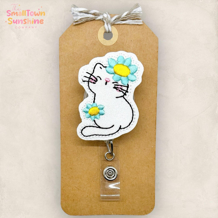 A white cat with a flower on its head is depicted on a brown tag or card, which is attached to a brown tag holder.