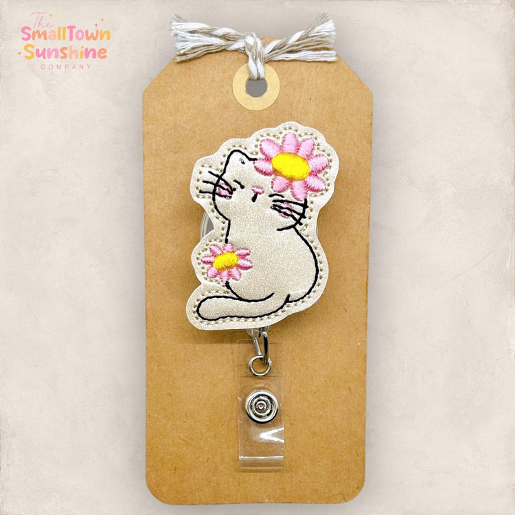 A badge with a cat embroidered on it, featuring a flower on its ear. The badge is attached to a brown tag with a clear plastic piece.