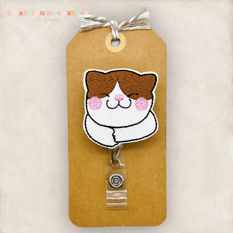 A brown and white cat embroidered badge with a clear plastic tag attached to it.