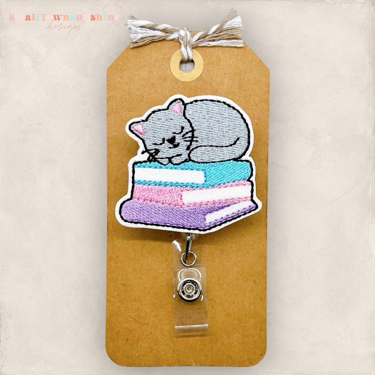 May include: A gray cat sleeping on a stack of colorful books. The cat is embroidered on a brown card with a clear plastic badge holder attached. The badge holder has a silver clip and a silver button.