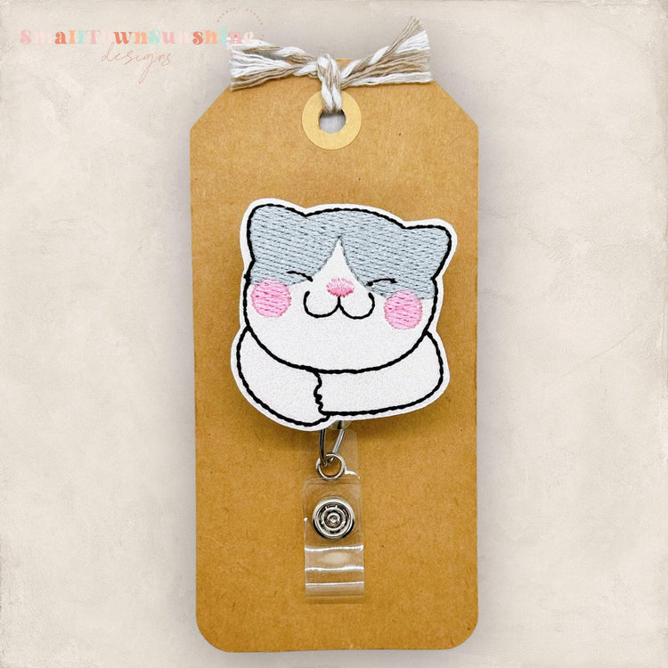 May include: A white and gray cat cartoon with pink cheeks, embroidered on a brown tag with a clear plastic retractable badge holder attached. The tag has the text 'Small Town Sue Shop Designs' at the top.
