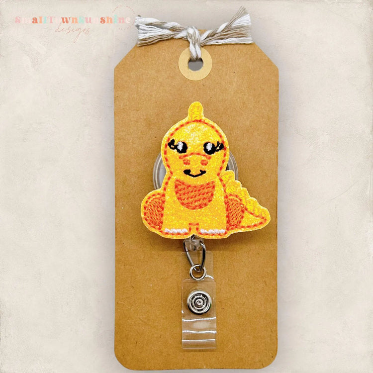 a brown paper tag with a yellow and orange embroidered dinosaur character attached to it, and a clear plastic clip on the tag.