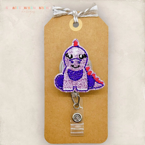 a brown paper tag with a purple and red embroidered dinosaur character attached to it, and a clear badge holder with a metal clip.