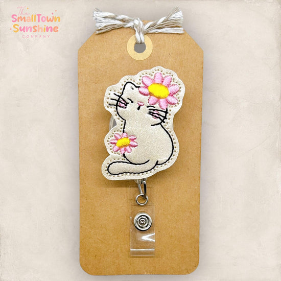 A badge with a cat embroidered on it, featuring a flower on its ear. The badge is attached to a brown tag with a clear plastic piece.