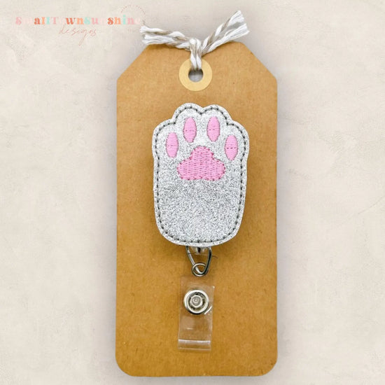A brown paper tag with a silver paw print embroidered on it, attached to a clear plastic clip.