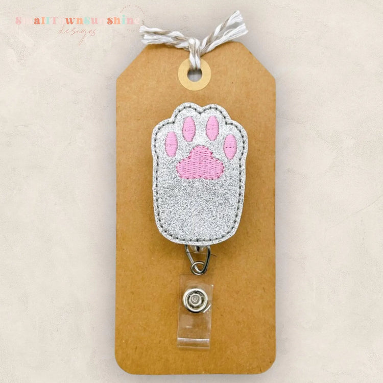 A brown paper tag with a silver paw print embroidered on it, attached to a clear plastic clip.