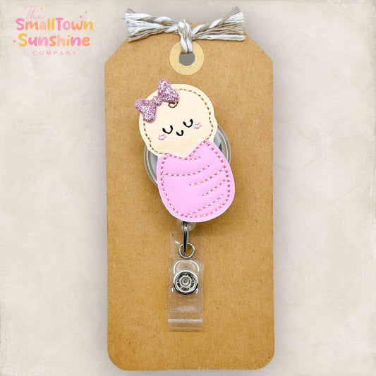 embroidered baby swaddled in a pink blanket with a pink glitter bow attached