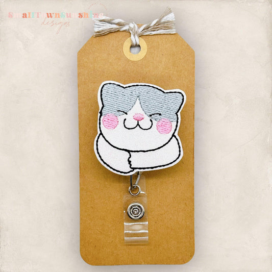 May include: A white and gray cat cartoon with pink cheeks, embroidered on a brown tag with a clear plastic retractable badge holder attached. The tag has the text &#39;Small Town Sue Shop Designs&#39; at the top.