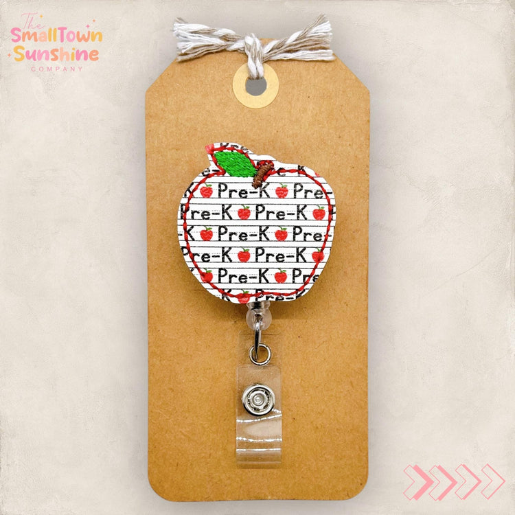 May include: An apple-shaped badge reel with the words "Pre-K" repeated on a white background with red apple accents. The reel is attached to a brown tag with a string and a clear plastic clip.